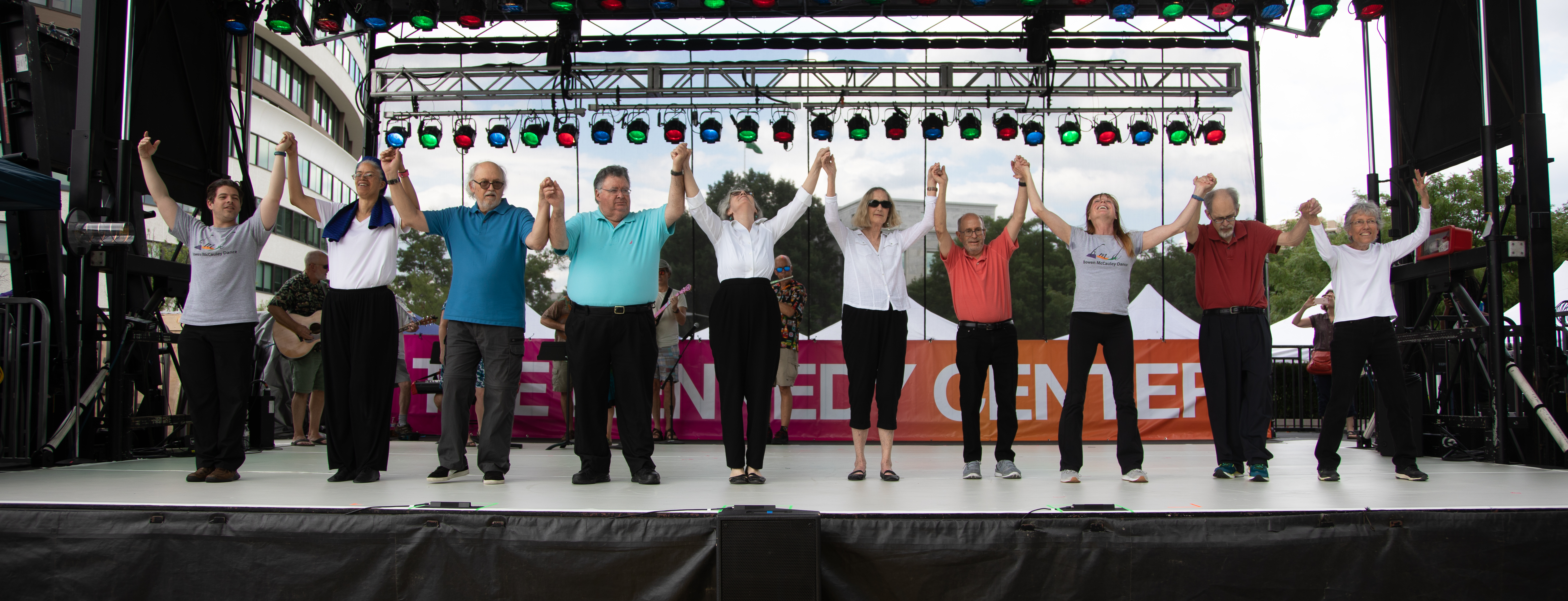Participants in the Dance for PD program stand in a row with their hands raised triumphantly on an outdoor stage at the Kennedy Center.